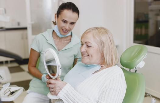 Starsza kobieta na fotelu dentystycznym uśmiech się widząc w lusterku swój nowy uśmiech